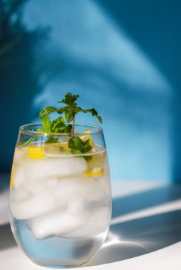 glass of water with mint, lemon and ice on white table, blue background