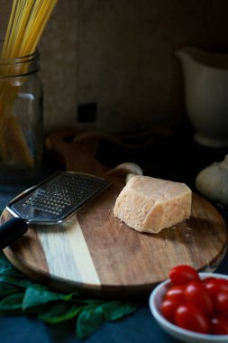 close up of parmesan cheese on wooden board with tomatoes, basil, pasta and grater