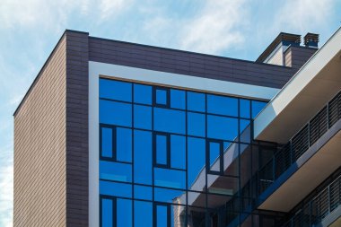 The top of the new building against the backdrop of the blue sky. Residential or office building with a facade of mirror windows. Brick wall and balconies with railing