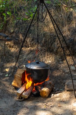 Cooking in a black tourist pot on an open fire. Cooking dinner over a campfire while camping outdoors. Cauldron with hot food is heated on burning wood