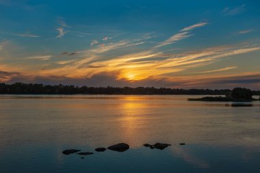 Beautiful sunset over the Dnieper River. Yellow cirrus clouds over the forest. Sun light reflection and stones in water