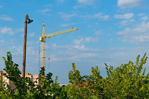 Construction crane with the flag of Ukraine and an old wooden pole. Blue sky and green foliage