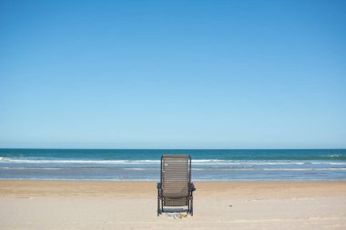 Arkasından kumsalda, deniz kıyısında boş bir güneş panjuru görünüyor.