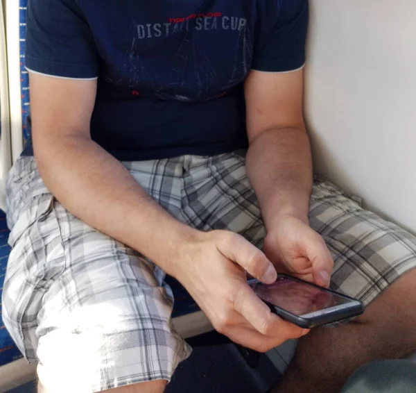 A man is looking at his smartphone in the subway. He\'s wearing checkered shorts. On a short sleeve t-shirt. The photo was taken while sitting.