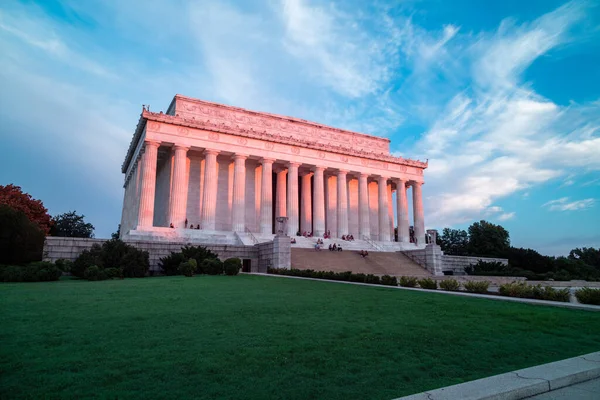 Pink light from sunrise illuminates the east-facing columns and exterior of the Lincoln Memorial in Washington, DC. Several sightseers sit on the marble steps.