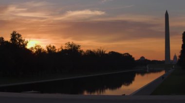 The Lincoln Memorial Reflecting Pool seen at sunrise on a summer morning. The Washington Monument and U.S. Capitol dome are seen in the distance. The sky is orange with dawn light. Unidentifiable pedestrians are seen.
