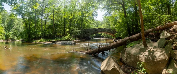 Washington 'daki Rock Creek Park' taki Boulder Köprüsü 'nün panoramik görüntüsü. Rock Creek ikonik köprünün altından akar; ön planda devrilmiş bir ağaç görülür..