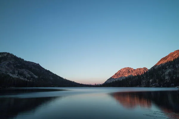 Toxaway Gölü, Idahos Sawtooth Wilderness 'da bir yaz akşamı gün batımında, güneş ışığının son ışıklarıyla kırmızımsı turuncu bir dağ sırtı ile görüldü. Su sahneyi yansıtıyor..