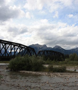 A black, metal railway bridge over a river flowing through the mountains.