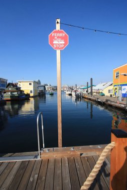An outside afternoon shot of a pier with a ferry stop sign surrounded by float homes in a harbour.