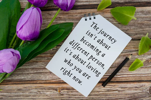 Motivational quote on notepad with flowers on wooden desk - The journey is not about becoming a different person, it is about loving who you are right now.
