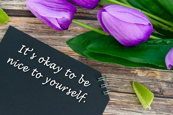 Motivational quote on black notepad with flowers on wooden desk - It is okay to be nice to yourself.