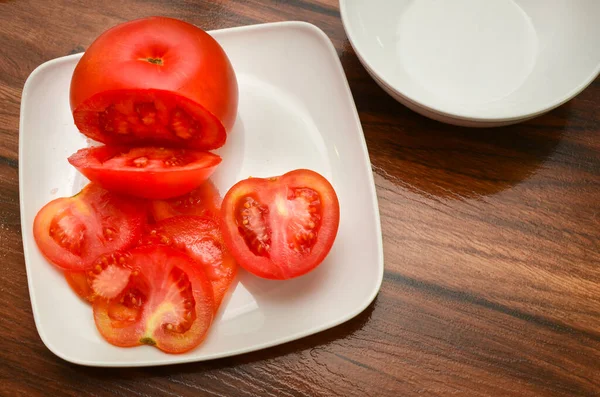 White plate seen from above with half a sliced tomato and several other slices on wooden table.