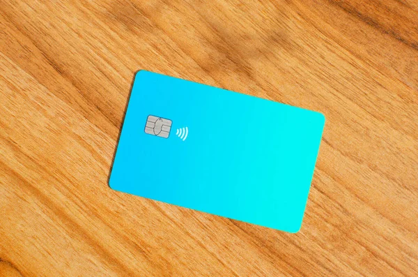 Plastic credit card with chip visible, on top of a table with soft lights and shadows. Blue card on wooden surface. Concept: finance, purchases, payments, loan, spending, investments and debts.