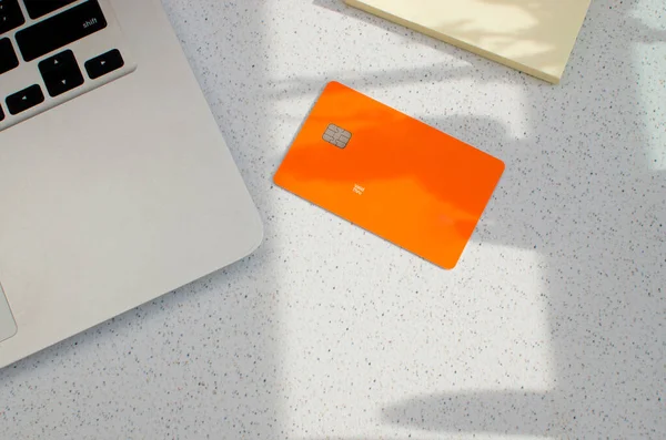 Kredi kartında görünür çip ve laptop var. Masanın üstünde yumuşak ışıklar ve gölgeler var. Beyaz yüzeyde turuncu renkli kart. Konsept: finans, satın almalar, ödemeler, kredi, harcama, yatırım ve borçlar.