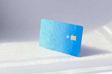 Yumuşak ışıklar ve gölgeli bir masanın üstünde görünen çipli plastik bir kredi kartı. Beyaz yüzeyde mavi kart. Konsept: finans, satın almalar, ödemeler, kredi, harcama, yatırım ve borçlar.