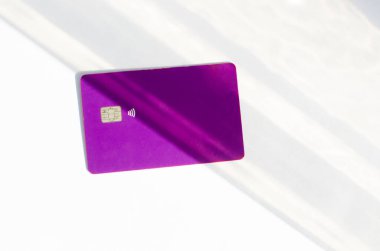 Yumuşak ışıklar ve gölgeli bir masanın üstünde görünen çipli plastik bir kredi kartı. Beyaz yüzeyde mor kart. Konsept: finans, satın almalar, ödemeler, kredi, harcama, yatırım ve borçlar.