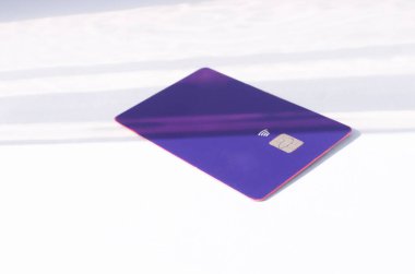 Yumuşak ışıklar ve gölgeli bir masanın üstünde görünen çipli plastik bir kredi kartı. Beyaz yüzeyde mor kart. Konsept: finans, satın almalar, ödemeler, kredi, harcama, yatırım ve borçlar.