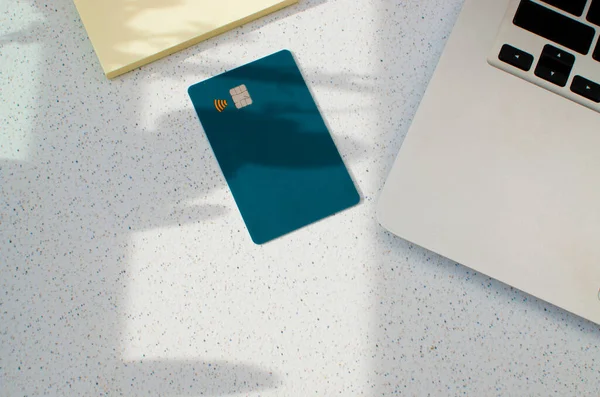 Kredi kartında görünür çip ve laptop var. Masanın üstünde yumuşak ışıklar ve gölgeler var. Beyaz yüzeyde yeşil renkli kart. Konsept: finans, satın almalar, ödemeler, kredi, harcama, yatırım ve borçlar.