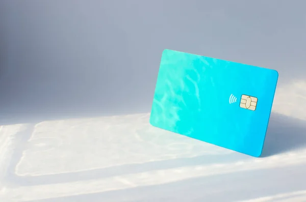 Plastic credit card with chip visible, on top of a table with soft lights and shadows. Blue card on white surface. Concept: finance, purchases, payments, loan, spending, investments and debts.