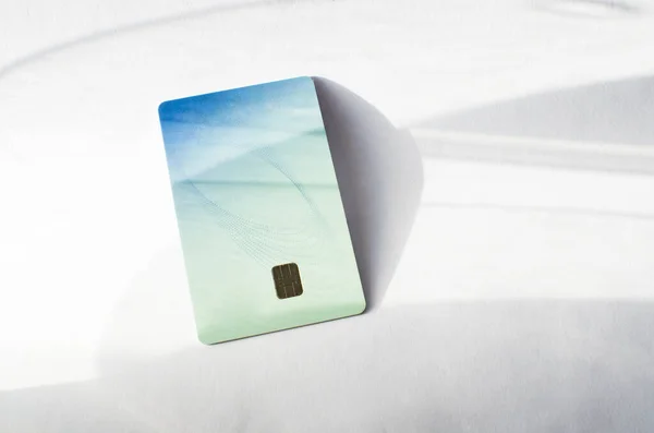 Plastic credit card with chip visible, on top of a table with soft lights and shadows. Blue and white color card on white surface. Concept: finances, purchases, payments, loans, spending and debts.