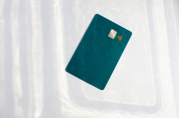 Plastic credit card with chip visible, on top of a table with soft lights and shadows. Green color card on white surface. Concept: finance, purchases, payments, loan, spending, investments and debts.