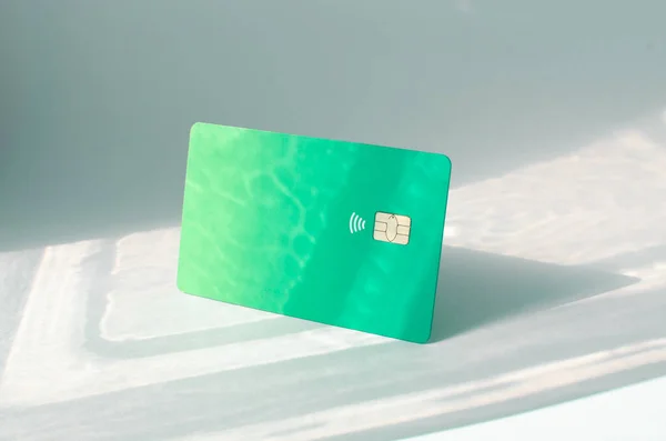 Plastic credit card with chip visible, on top of a table with soft lights and shadows. Green color card on white surface. Concept: finance, purchases, payments, loan, spending, investments and debts.
