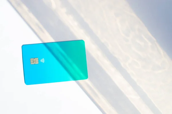 Plastic credit card with chip visible, on top of a table with soft lights and shadows. Blue card on white surface. Concept: finance, purchases, payments, loan, spending, investments and debts.