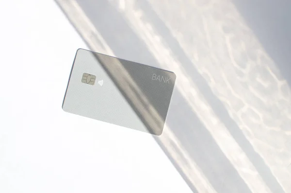 Plastic credit card with chip visible, on top of a table with soft lights and shadows. Gray card on white surface. Concept: finance, purchases, payments, loan, spending, investments and debts.