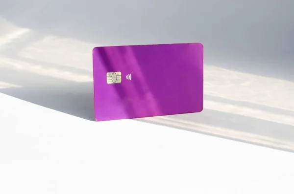 Plastic credit card with chip visible, on top of a table with soft lights and shadows. Purple card on white surface. Concept: finance, purchases, payments, loan, spending, investments and debts.