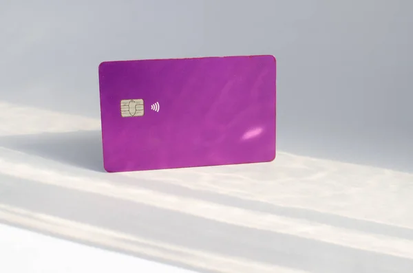 Plastic credit card with chip visible, on top of a table with soft lights and shadows. Purple card on white surface. Concept: finance, purchases, payments, loan, spending, investments and debts.
