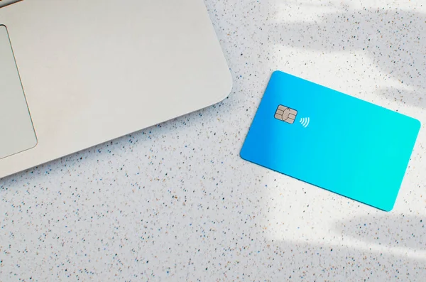 Plastic credit card with chip visible, on top of a table with soft lights and shadows. Blue card on white surface. Concept: finance, purchases, payments, loan, spending, investments and debts.