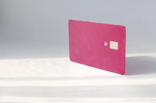 Plastic credit card with chip visible, on top of a table with soft lights and shadows. Red color card on white surface. Concept: finance, purchases, payments, loan, spending, investments and debts.