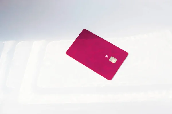 Plastic credit card with chip visible, on top of a table with soft lights and shadows. Red color card on white surface. Concept: finance, purchases, payments, loan, spending, investments and debts.