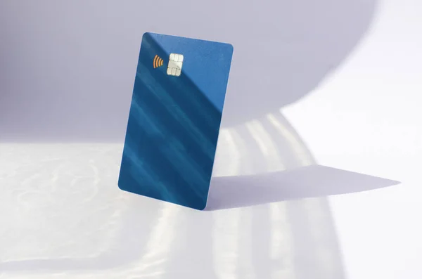 Plastic credit card with chip visible, on top of a table with soft lights and shadows. Blue card on white surface. Concept: finance, purchases, payments, loan, spending, investments and debts.