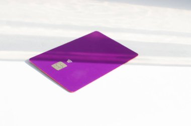 Yumuşak ışıklar ve gölgeli bir masanın üstünde görünen çipli plastik bir kredi kartı. Beyaz yüzeyde mor kart. Konsept: finans, satın almalar, ödemeler, kredi, harcama, yatırım ve borçlar.