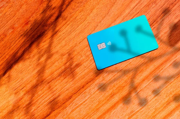Plastic credit card with chip visible, on top of a table with soft lights and shadows. Blue card on wooden surface. Concept: finance, purchases, payments, loan, spending, investments and debts.