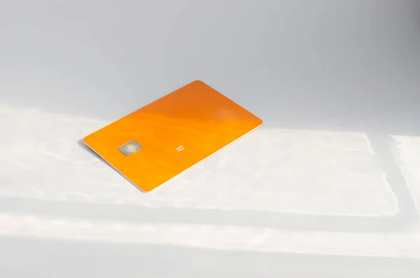 Plastic credit card with chip visible, on top of a table with soft lights and shadows. Orange color card on white surface. Concept: finance, purchases, payments, loan, spending, investments and debts.