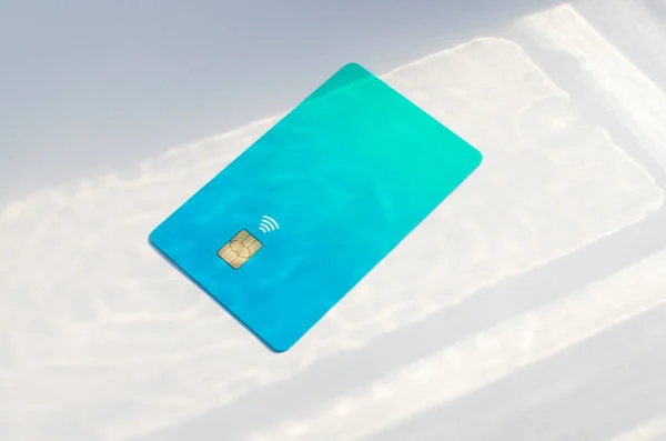 Plastic credit card with chip visible, on top of a table with soft lights and shadows. Blue card on white surface. Concept: finance, purchases, payments, loan, spending, investments and debts.