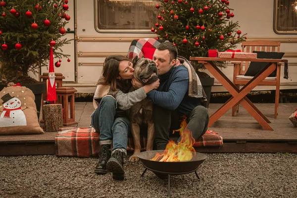 Happy couple in love and their dog spend time together on Christmas holidays. Cane Corso and his owners are celebrating the new year in the open air near the motor home and the fire. New Year