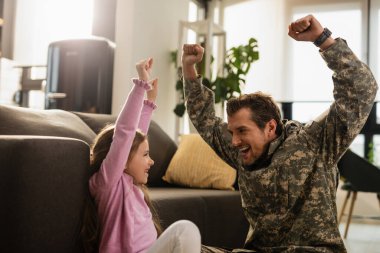 Happy army soldier and his small daughter having fun while celebrating reunion with arms raised in the living room. 