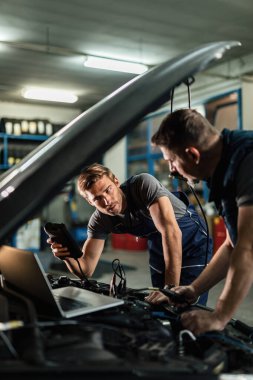 Young car repairman talking with his coworker while checking engine performance with diagnostic tools in auto repair shop. 
