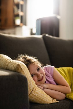 Happy small girl resting on the sofa in the living room and looking at camera. 