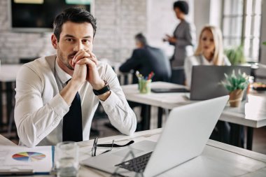 Young pensive businessman working at desk in the office and thinking of something. There are people in the background. 