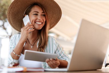 Young smiling woman sitting in a cafe and talking on mobile phone while using touchpad and laptop. 