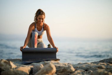 Happy female athlete getting ready for sports training and preparing her exercises mat on a rock at the beach. Copy space. 