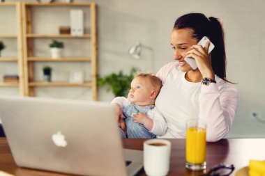Young happy woman communicating over mobile phone while relaxing with her baby at home. 