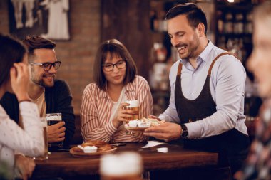 Young happy waiter serving a snack to customers who are drinking beer in a pub. 