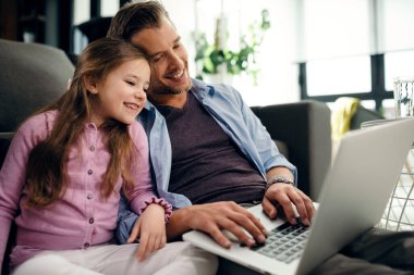 Happy father and daughter sitting close to each other while using computer and surfing the internet at home. 