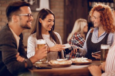 Happy woman paying to a waitress via contactless smart phone payment in a bar. 